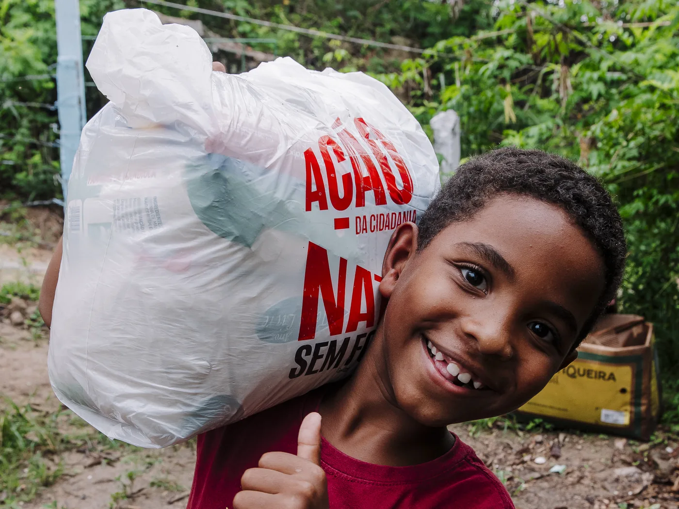 Menino sorrindo com o polegar erguido, carregando no ombro uma sacola de alimentos da Ação da Cidadania